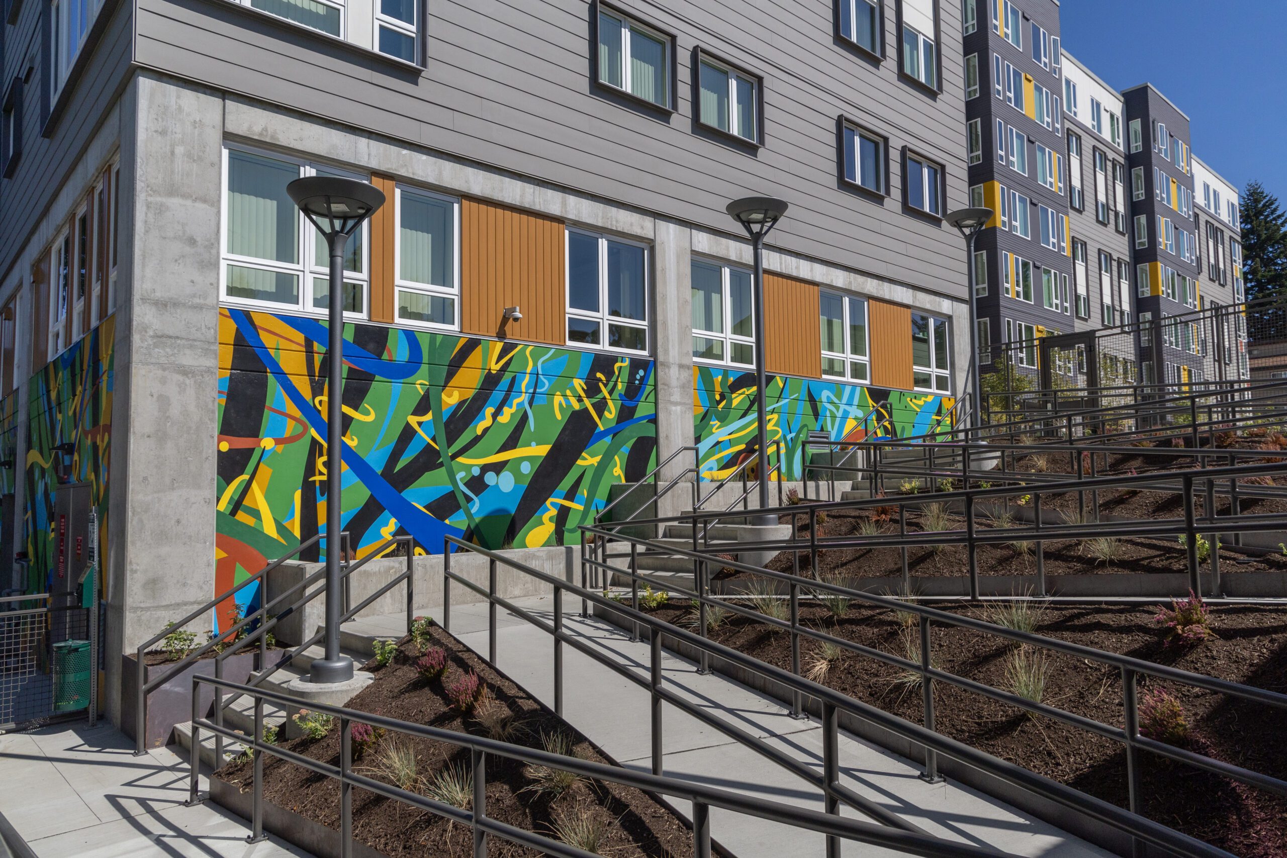 An exterior view of a modern, multi-story residential building with gray and yellow paneling. A vibrant, abstract mural with blue, green, and yellow patterns decorates the lower portion of the building. A series of accessibility ramps with black metal railings zigzags up a landscaped hillside, providing an inclusive pathway to the entrance.